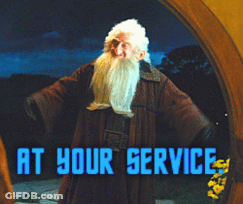A Man With A Beard Stands In Front Of A Sign That Reads At Your Service GIF