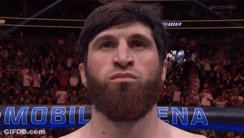 A Man With A Beard Stands In Front Of A Sign That Says Mobil Ena GIF