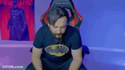 A Man With A Beard Wearing A Batman Shirt Is Sitting In A Chair GIF