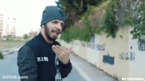 A Man With A Beard Wearing A Beanie And A Black Jacket Is Standing In Front Of A Wall . GIF