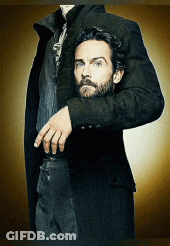 A Man With A Beard Wearing A Black Coat Has His Hand On His Pocket GIF