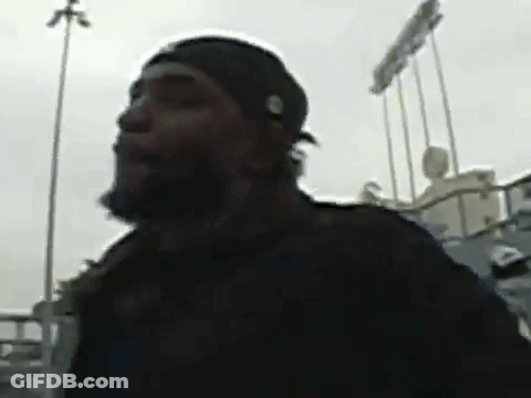 A Man With A Beard Wearing A Black Hat Is Standing In Front Of A Baseball Stadium . GIF