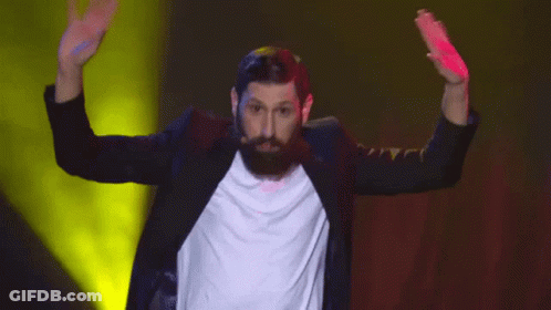 A Man With A Beard Wearing A Black Jacket And A White Shirt Is Dancing On A Stage GIF