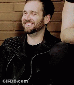 A Man With A Beard Wearing A Black Leather Jacket Is Smiling While Sitting In Front Of A Brick Wall . GIF