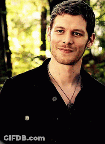 A Man With A Beard Wearing A Black Shirt And A Necklace Smiles For The Camera GIF
