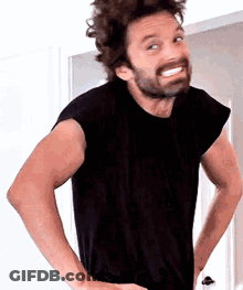 A Man With A Beard Wearing A Black Shirt Is Standing With His Hands On His Hips And Smiling . GIF