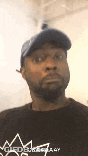 A Man With A Beard Wearing A Blue Hat And A Black Shirt With A Crown On It . GIF