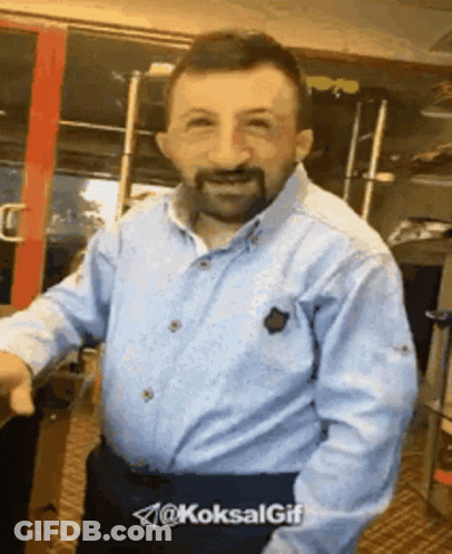 A Man With A Beard Wearing A Blue Shirt Is Standing In Front Of A Red Door .. GIF