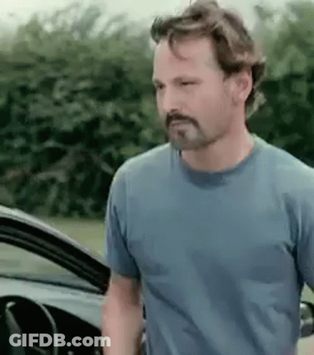 A Man With A Beard Wearing A Blue Shirt Is Standing Next To A Car . GIF