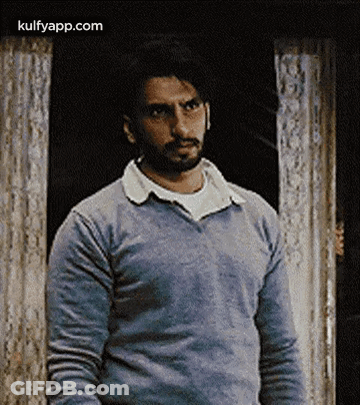 A Man With A Beard Wearing A Blue Sweater Is Standing In Front Of A Door . GIF