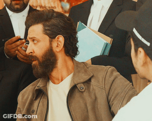 A Man With A Beard Wearing A Brown Jacket Is Talking To Another Man GIF