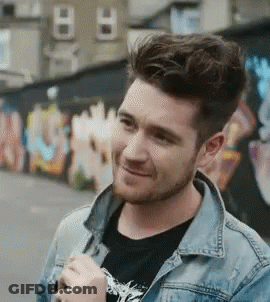 A Man With A Beard Wearing A Denim Jacket And A Black Shirt Is Smiling In Front Of A Graffiti Wall . GIF