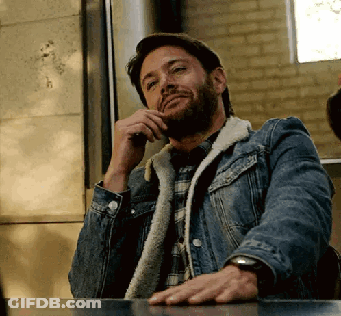 A Man With A Beard Wearing A Denim Jacket Is Sitting At A Table Talking On A Cell Phone . GIF