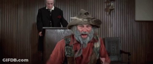 A Man With A Beard Wearing A Feathered Hat Stands In Front Of A Judge GIF