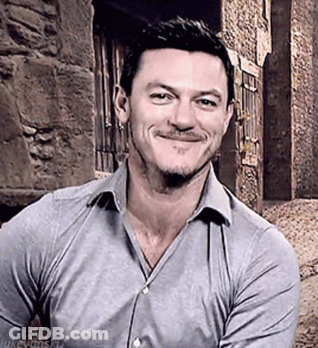 A Man With A Beard Wearing A Grey Shirt Is Smiling In Front Of A Stone Wall GIF