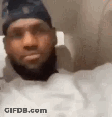 A Man With A Beard Wearing A Hat And A White Shirt Is Sitting In A Chair . GIF