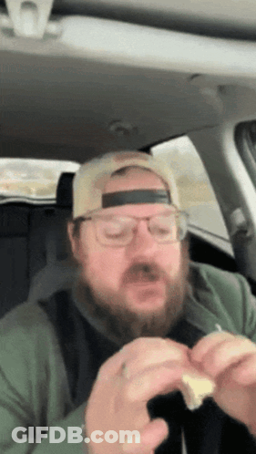A Man With A Beard Wearing A Hat And Glasses Is Eating A Sandwich GIF
