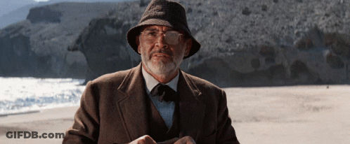 A Man With A Beard Wearing A Hat And Glasses Stands On A Beach GIF