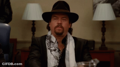 A Man With A Beard Wearing A Hat And Scarf Is Sitting At A Table GIF