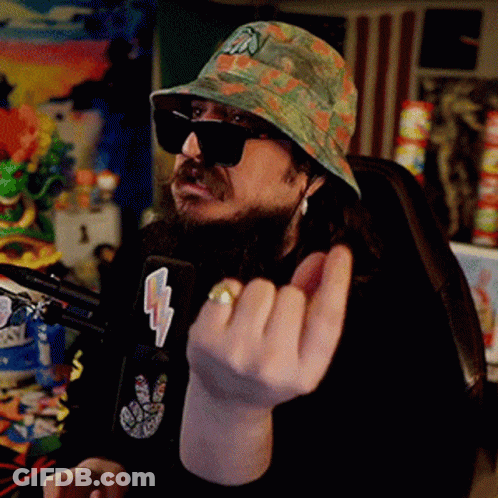 A Man With A Beard Wearing A Hat And Sunglasses Is Sitting In Front Of A Microphone Giving The Middle Finger . GIF