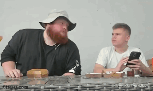 A Man With A Beard Wearing A Hat Is Sitting At A Table With Another Man . GIF