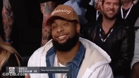 A Man With A Beard Wearing A Hat Is Smiling While Sitting In A Crowd . GIF