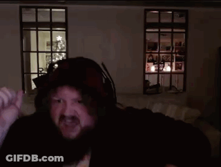 A Man With A Beard Wearing A Hooded Jacket Is Standing In A Dark Room With A Christmas Tree In The Background GIF