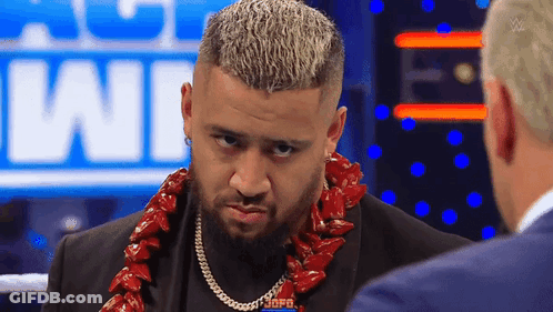 A Man With A Beard Wearing A Lei Is Talking To Another Man In A Suit . GIF