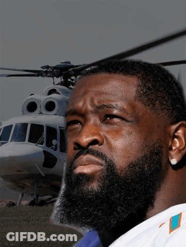 A Man With A Beard Wearing A Miami Dolphins Shirt Stands In Front Of A Helicopter GIF