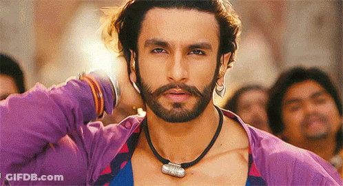 A Man With A Beard Wearing A Purple Shirt And A Black Necklace Is Standing In Front Of A Crowd . GIF