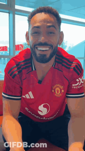 A Man With A Beard Wearing A Red Adidas Jersey Is Smiling While Sitting Down . GIF