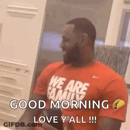 A Man With A Beard Wearing A Red Shirt That Says `` We Are Family '' Is Sitting In Front Of A Window . GIF