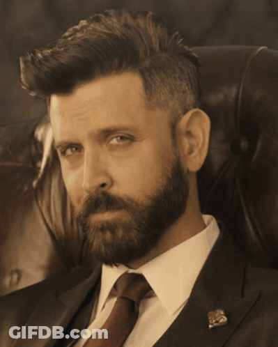A Man With A Beard Wearing A Suit And Tie Is Sitting In A Chair GIF