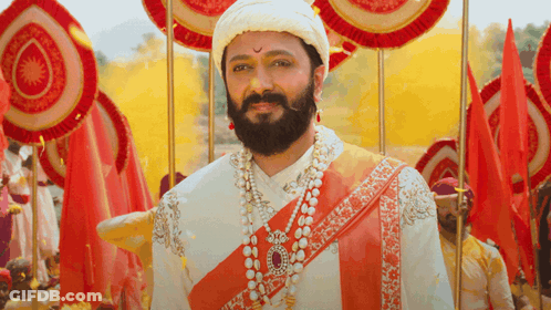A Man With A Beard Wearing A Turban And Pearls Stands In Front Of Red Flags GIF