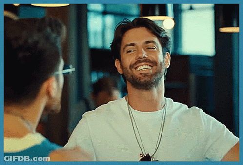 A Man With A Beard Wearing A White T-Shirt Smiles While Talking To Another Man GIF