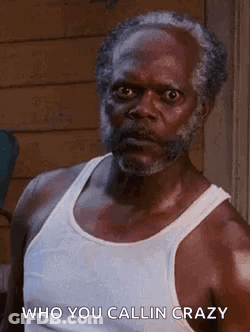 A Man With A Beard Wearing A White Tank Top Is Standing In Front Of A Building . GIF