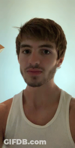 A Man With A Beard Wearing A White Tank Top Looks At The Camera GIF