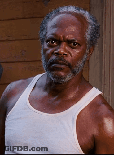 A Man With A Beard Wearing A White Tank Top Looks At The Camera GIF