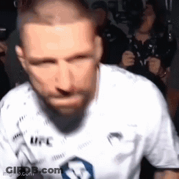 A Man With A Beard Wearing A White UFC Shirt Is Standing In Front Of A Crowd . GIF