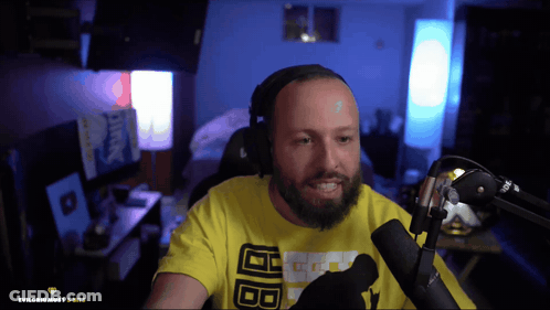 A Man With A Beard Wearing A Yellow Shirt With The Letters Bb On It Is Sitting In Front Of A Microphone GIF