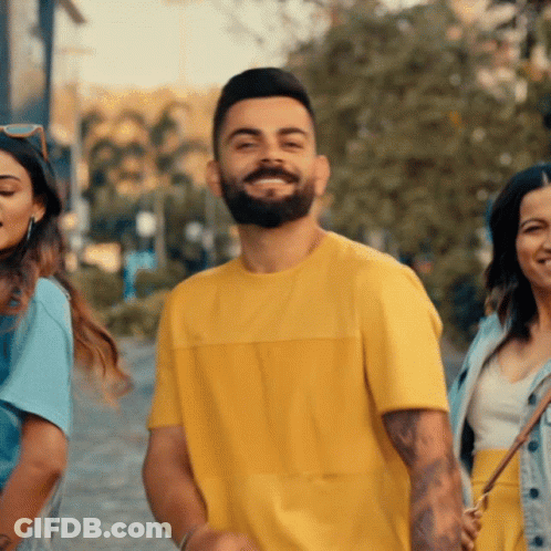 A Man With A Beard Wearing A Yellow T-Shirt Smiles With Two Women Behind Him GIF