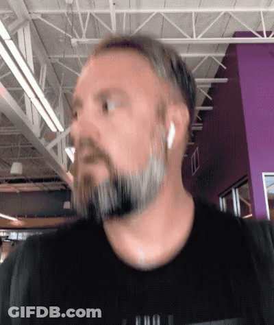 A Man With A Beard Wearing Ear Buds And A Black Shirt With A Purple Wall In The Background GIF