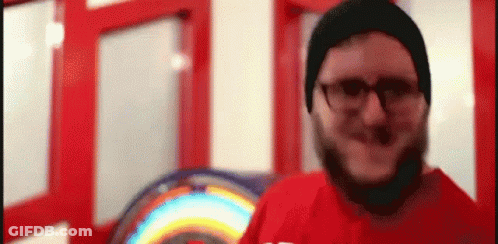 A Man With A Beard Wearing Glasses And A Beanie Is Standing In Front Of A Rainbow Sign . GIF