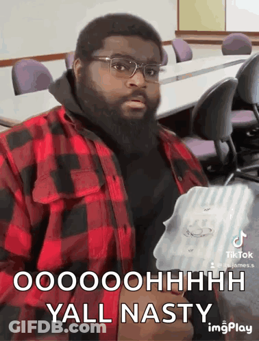 A Man With A Beard Wearing Glasses And A Plaid Shirt Is Sitting In A Classroom Holding A Box . GIF
