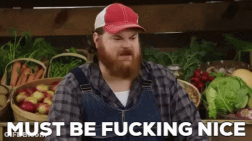 A Man With A Beard Wearing Overalls And A Hat Is Standing In Front Of Baskets Of Fruit And Vegetables . GIF