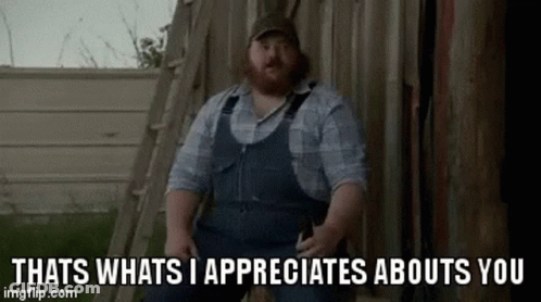 A Man With A Beard Wearing Overalls Is Sitting In Front Of A Wooden Shed . GIF