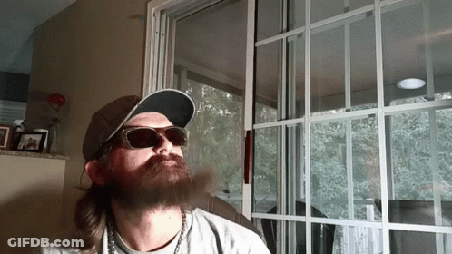 A Man With A Beard Wearing Sunglasses And A Hat Is Smoking A Cigarette GIF
