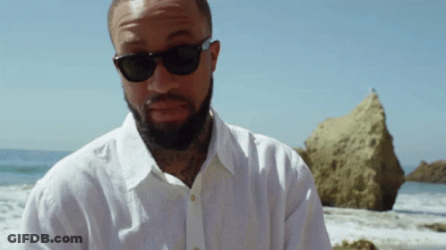 A Man With A Beard Wearing Sunglasses And A White Shirt Stands On The Beach GIF