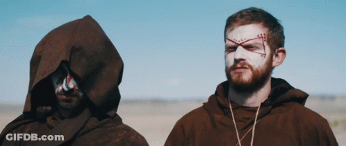 A Man With A Cross Painted On His Face Stands Next To Another Man With A Hood GIF