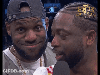 A Man With A Crown On His Head Talks Into A Microphone Next To Another Man GIF